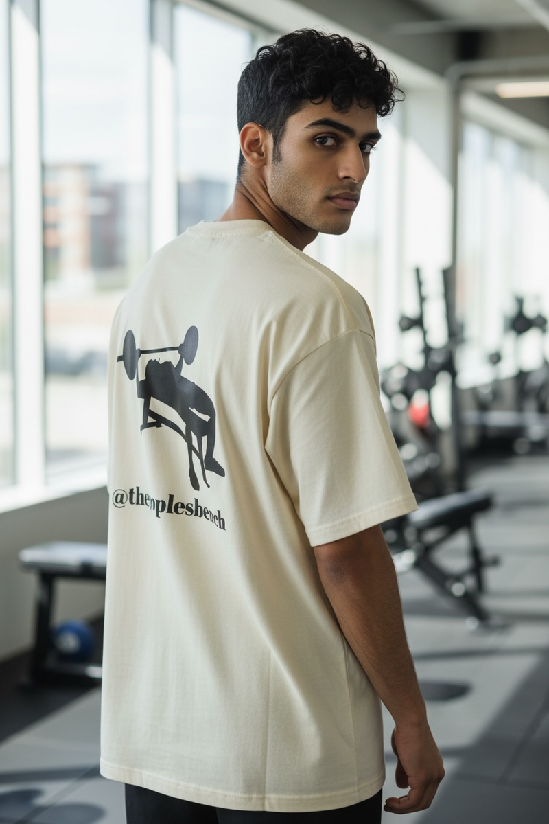 A man wears a cream t-shirt with a black graphic of a person lifting weights and text.